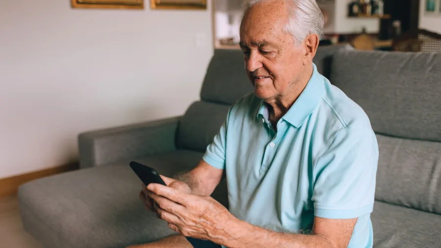 Älterer Mann sitzt auf dem Sofa und prüft mit dem Smartphone seine Unterlagen zur Pflegekasse